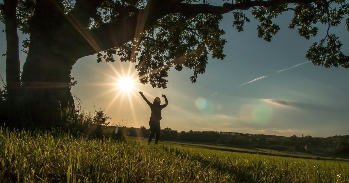 Person joyfully raising arms under a large tree at sunset in a grassy field, symbolizing hope, peace, and renewal.